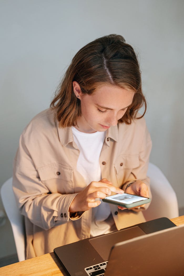 Focused young woman using a smartphone while working on a laptop indoors.